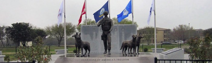 Image of the Military Working Dog Teams National Monument. The monument features a bronze handler and four canines standing atop a pedestal that says “Guardians of America’s Freedom”.
